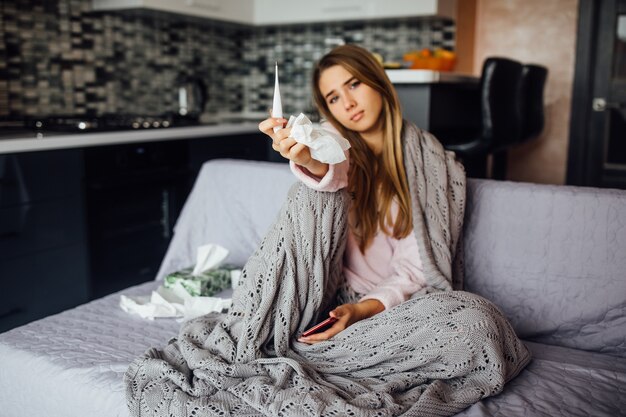 Junge Frau ruht im Bett und bedeckt die Nase mit einem Taschentuch, das ein Thermometer hält