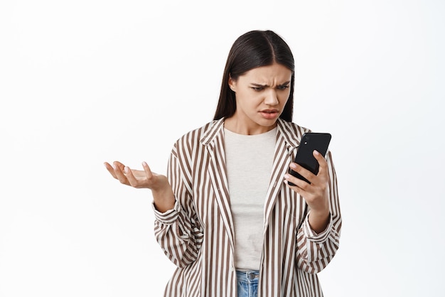 Junge Frau schaut verwirrt auf ihr Telefon, breitet die Hand seitwärts aus und verzieht das Gesicht, als sie eine seltsame Nachricht auf dem Smartphone liest, das vor weißem Hintergrund steht