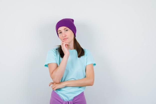 Junge Frau schiefes Kinn zur Hand in blauem T-Shirt, lila Mütze und sieht ernst aus, Vorderansicht.