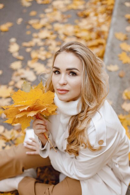 Junge Frau sitzt auf einem Bordstein im herbstlichen Wald. Blonde Frau, die gelbe Blätter hält. Mädchen mit beigem Mantel und brauner Hose.