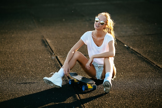 Kostenloses Foto junge frau sitzt auf einem skateboard mit ihrer sonnenbrille an während des sonnenuntergangs.