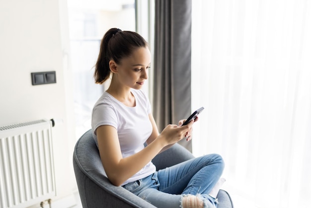 Junge Frau sitzt im Stuhl mit Smartphone trägt lässige Kleidung im Haus drinnen