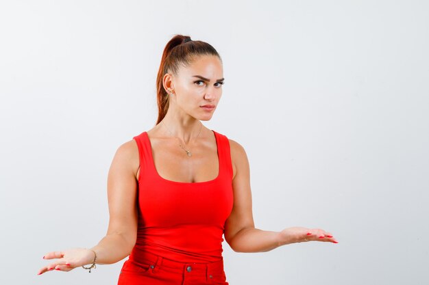 Junge Frau spreizt Handflächen beiseite im roten Trägershirt, in der Hose und in der ernsthaften Vorderansicht.