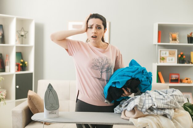 Junge Frau steht hinter Bügelbrett mit Kleidung im Wohnzimmer