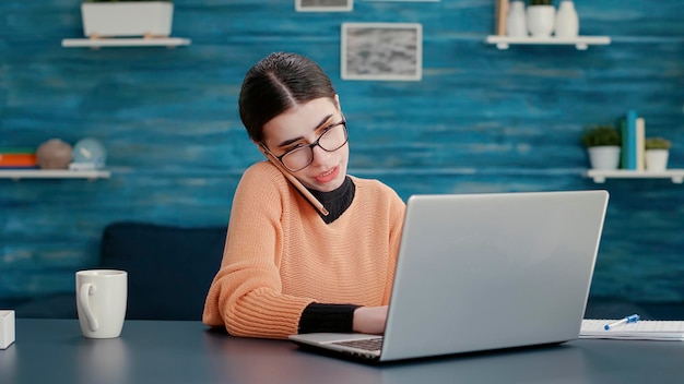 Junge Frau telefoniert und benutzt Laptop am Schreibtisch zu Hause, nimmt an Online-Klassenunterricht für Fernunterricht teil. Moderne Kommunikation auf dem Smartphone, Schularbeiten am Computer.