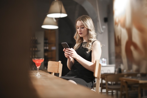 junge frau trinkt einen cocktail in einer bar und liest von ihrem smartphone