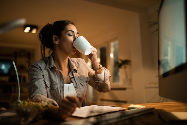 Junge Frau trinkt Kaffee mit geschlossenen Augen, während sie nachts zu Hause lernt