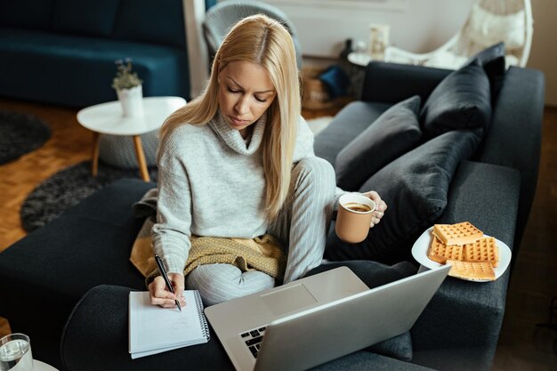 Junge Frau trinkt Kaffee, während sie an einem Computer arbeitet und sich im Wohnzimmer Notizen macht