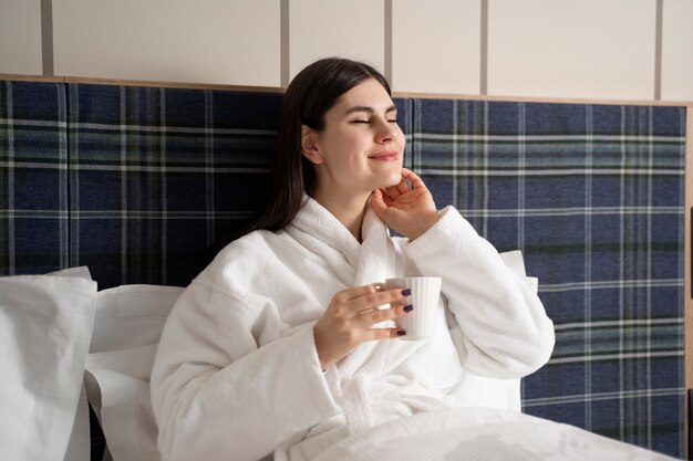 Junge Frau trinkt Kaffee, während sie auf dem Bett in einem Hotelzimmer sitzt