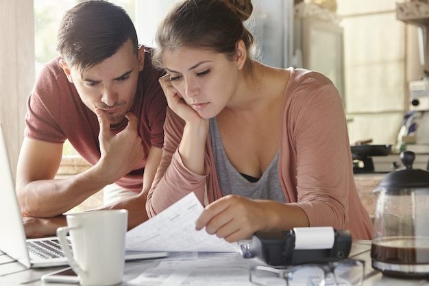 Junge Frau und ihr arbeitsloser Ehemann mit vielen Schulden, die gemeinsam Papierkram in der Küche erledigen