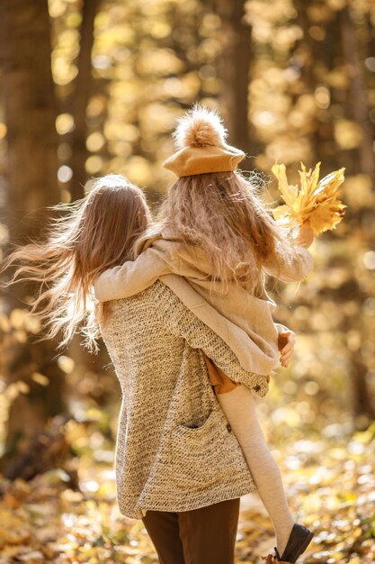 Junge Frau und kleines Mädchen im Herbstwald. Frau, die ihre Tochter auf ihren Händen hält. Mädchen, das modische Kleidung trägt und gelbe Blätter hält.