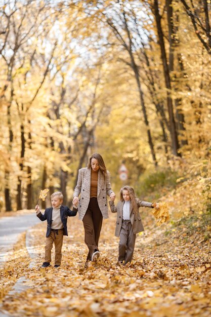 Junge Frau und kleines Mädchen und Junge, die im Herbstwald spazieren. Frau, ihre Tochter und ihr Sohn spielen und Spaß haben. Mädchen, das graues Kostüm der Mode und blaue Jacke des Jungen trägt.