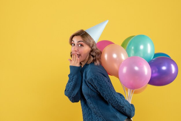 junge Frau versteckt bunte Luftballons hinter ihrem Rücken auf gelb