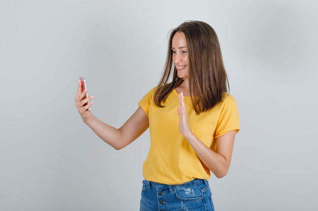 Junge Frau winkt Hand auf Videoanruf in T-Shirt, Shorts und schaut froh