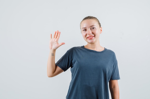 Junge Frau winkt Hand, um sich im grauen T-Shirt zu verabschieden und froh zu schauen