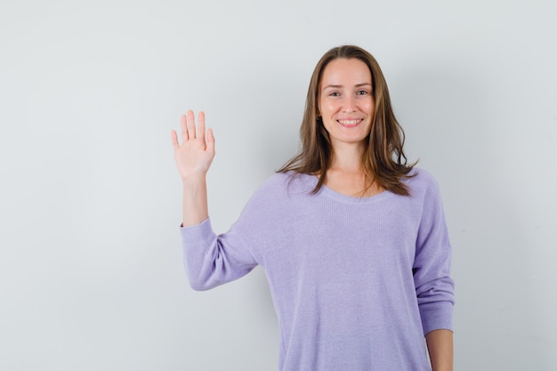Junge Frau winkt Hand zum Begrüßen in lila Bluse und schaut froh