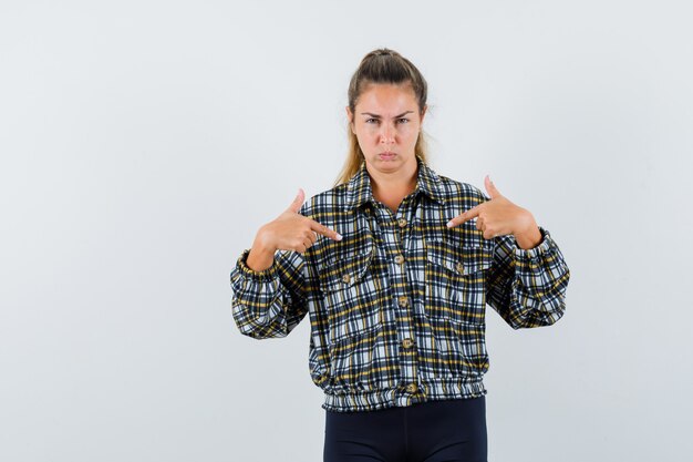 Junge Frau zeigt auf sich selbst in Hemd, Shorts und schaut zweifelhaft, Vorderansicht.