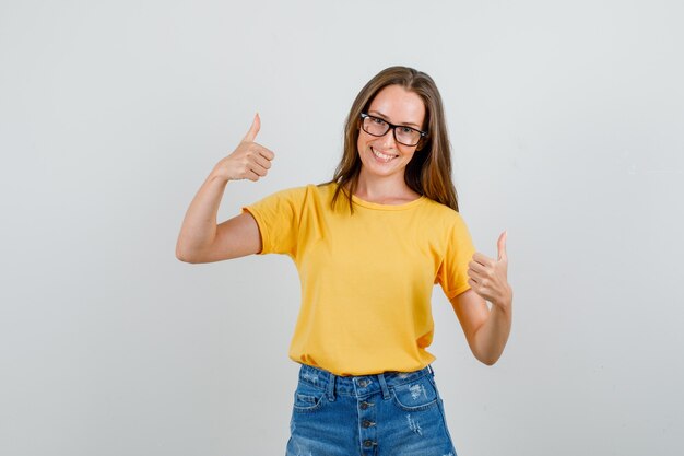 Junge Frau zeigt Daumen hoch und lächelt in T-Shirt, Shorts, Brille Vorderansicht.