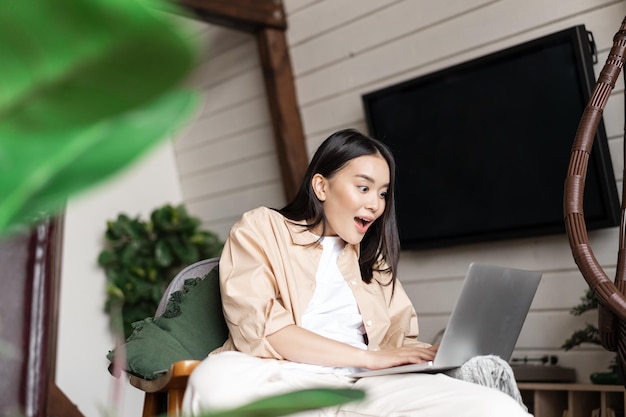 Junge Frau zu Hause, die ihren Laptop mit erstauntem Wow-Gesicht betrachtet, schockiert über Ankündigungsbenachrichtigung...