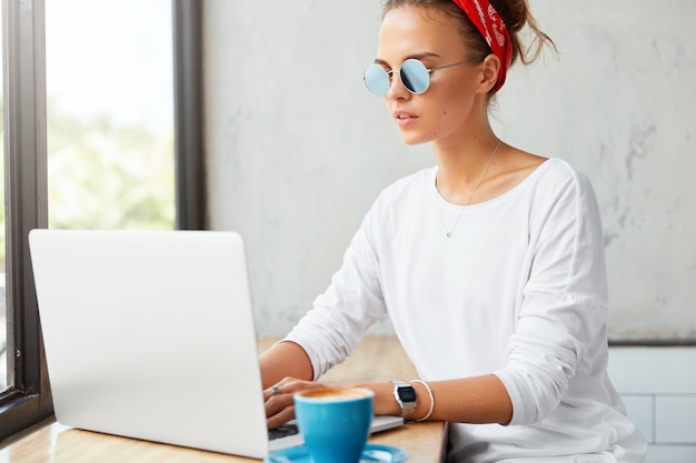 Junge freiberuflerin arbeitet remote am laptop im café, gibt tastaturinformationen aus und trinkt kaffee. frau chattet mit freunden in sozialen netzwerken, verbunden mit drahtlosem internet im restaurant