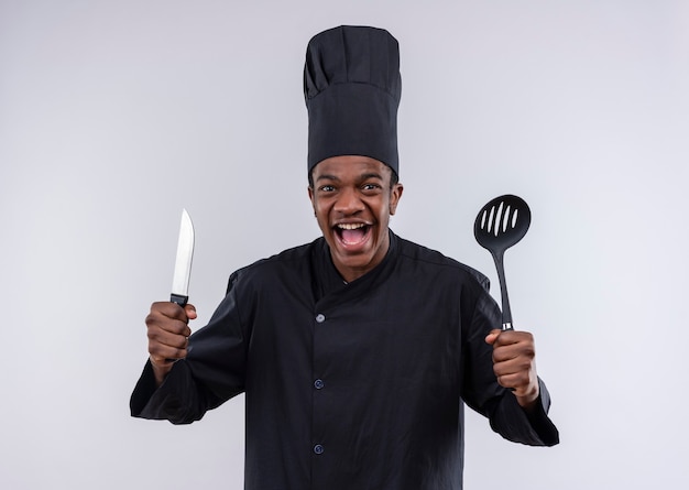 Junge freudige afroamerikanische Köchin in der Kochuniform hält Messer und Spatel isoliert auf weißer Wand