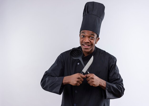 Junge freudige afroamerikanische Köchin in der Kochuniform hält Messer und Spatel lokalisiert auf weißer Wand