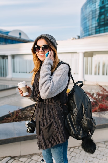 Junge freudige Frau mit Kaffee, zum des Gehens am sonnigen kalten Tag in der Großstadt zu gehen. Hübsche Frau, die warmen Winterwollpullover, moderne Sonnenbrille trägt, am Telefon spricht, mit Kamera und Tasche reist, glücklich.