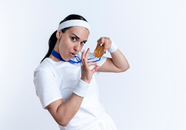 Kostenloses Foto junge freudige kaukasische sportliche frau, die stirnband und armbänder trägt, hält goldmedaille und gestikuliert ok handzeichen lokalisiert auf weißem raum mit kopienraum