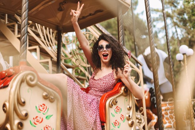 Junge fröhliche Dame mit dunklem lockigem Haar in Sonnenbrille und Kleid, die Lolli-Pop-Süßigkeiten in der Hand hält und zwei Fingergesten zeigt, während sie auf Karussell im Vergnügungspark fährt