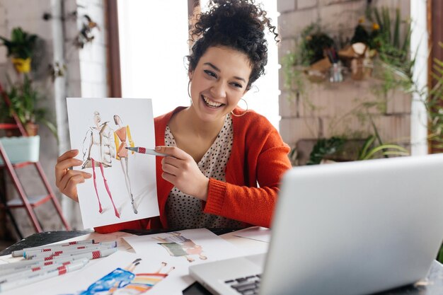 Junge fröhliche Frau mit dunklem, lockigem Haar, die glücklich am Tisch sitzt und Modeillustrationen auf dem Laptop zeigt, während sie Zeit in einer modernen, gemütlichen Werkstatt mit großen Fenstern verbringt