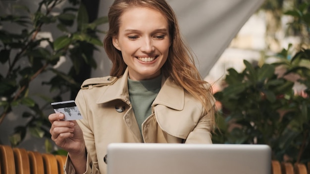 Junge, fröhliche Frau mit Kreditkarte, die ihr Bankkonto auf dem Laptop über die kostenlose Internetverbindung im Straßencafé überprüft. Modernes Technologiekonzept