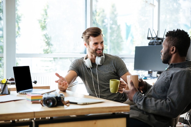 Junge fröhliche Kollegen, die im Büro sitzen und miteinander sprechen