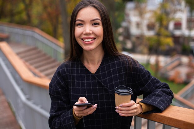 Junge, fröhliche, lässige Frau im Mantel mit Kaffee zum Mitnehmen und Handy, die glücklich in die Kamera im Stadtpark schaut