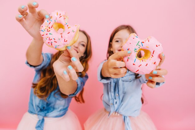 Junge fröhliche Mutter und süße lächelnde Tochter, die Spaß mit leckeren Donuts haben, die auf Teeparty mit Familie warten. Kleines Mädchen mit ihrer Mutter zeigt Donuts, die sie zusammen gekocht haben und lachen