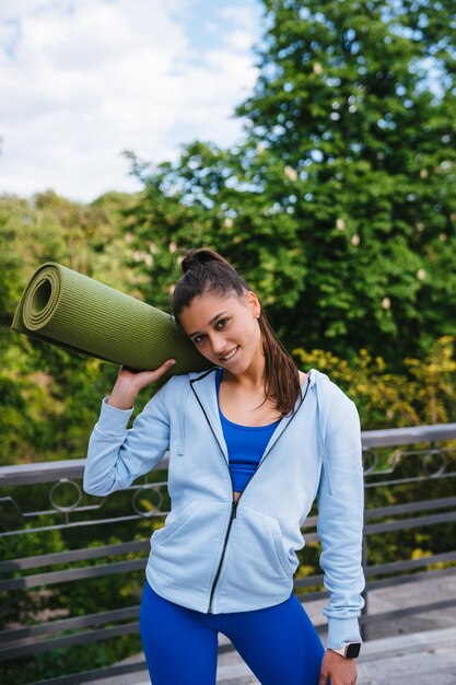 Junge fröhliche Sportfrau, die im Stadtpark hält Fitness-Teppich geht.