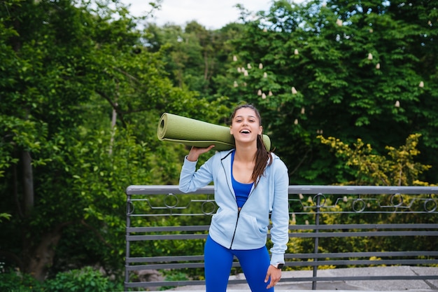 Junge fröhliche Sportfrau, die im Stadtpark hält Fitness-Teppich geht.