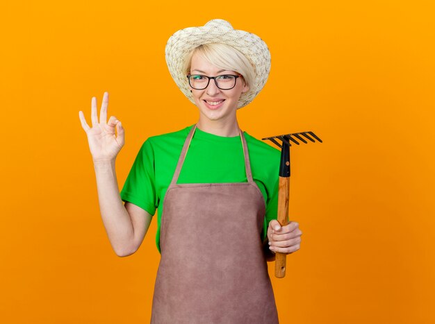 Junge Gärtnerfrau mit kurzen Haaren in der Schürze und im Hut, die Mini-Rechen halten, der Kamera lächelnd zeigt, ok Zeichen zeigend, das über orange Hintergrund steht