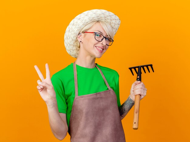 Junge Gärtnerfrau mit kurzen Haaren in der Schürze und im Hut, die Mini-Rechen-Lookign an der lächelnden Kamera halten, die V-Zeichen zeigt, das über orange Hintergrund steht