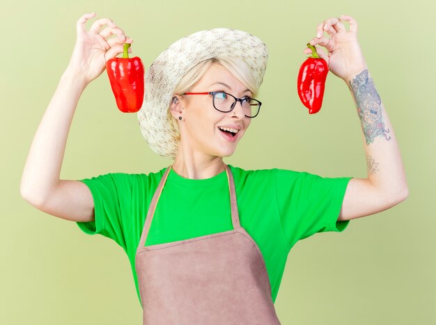 Junge Gärtnerfrau mit kurzen Haaren in der Schürze und im Hut, die rote Paprika halten, die Spaß haben, fröhlich stehend über hellem Hintergrund zu lächeln