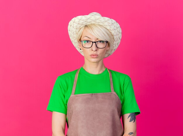 Junge Gärtnerfrau mit kurzen Haaren in Schürze und Hut, die Kamera mit ernstem Gesicht betrachten