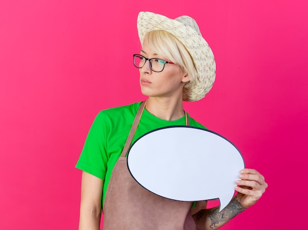 Junge Gärtnerfrau mit kurzen Haaren in Schürze und Hut, die leere Sprechblase halten