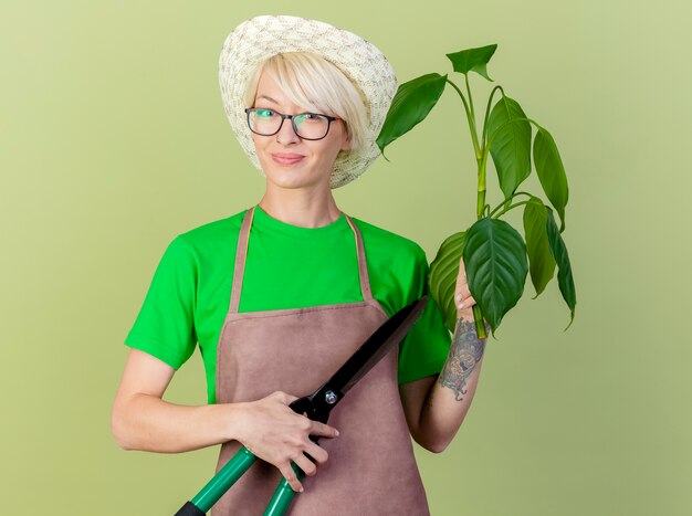 Junge Gärtnerin mit kurzen Haaren in der Schürze und im Hut, die Pflanze und Heckenscheren halten, die Kamera mit Lächeln auf Gesicht betrachten, das über hellem Hintergrund steht