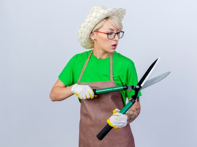 Kostenloses Foto junge gärtnerin mit kurzen haaren in schürze und hut, die eine heckenschere hält und sie überrascht anschaut