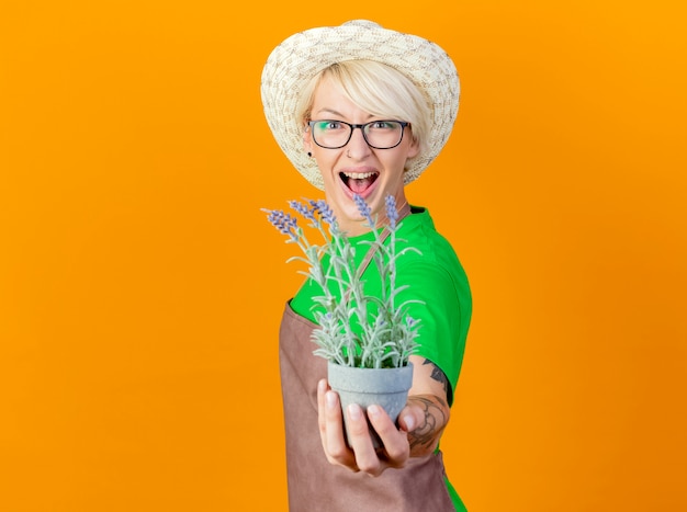 Junge gärtnerin mit kurzen haaren in schürze und hut, die eine topfpflanze zeigt, die mit glücklichem gesicht auf orangefarbenem hintergrund lächelt