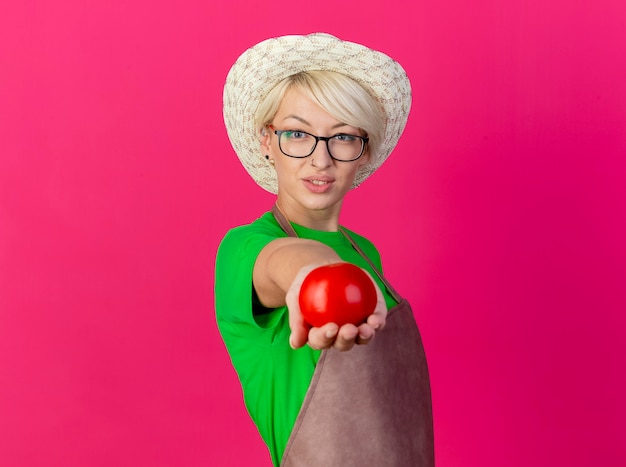 Kostenloses Foto junge gärtnerin mit kurzen haaren in schürze und hut, die frische tomate anbietet