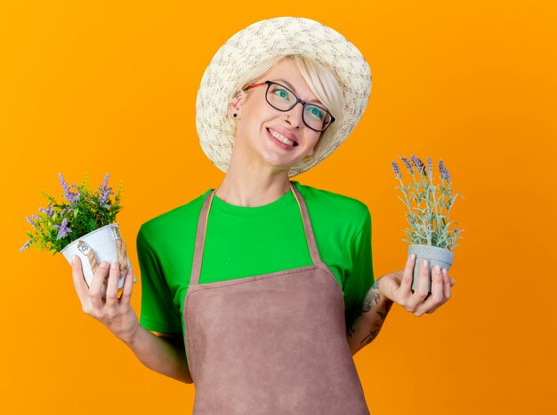 Junge Gärtnerin mit kurzen Haaren in Schürze und Hut, die Topfpflanzen halten, schauen beiseite lächelnd mit glücklichem Gesicht, das über orange Hintergrund steht