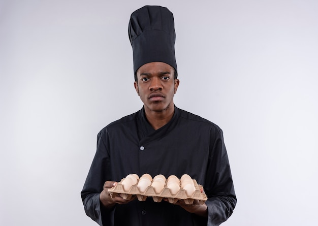 Junge genervte afroamerikanische Köchin in der Kochuniform hält Charge von Eiern lokalisiert auf weißem Hintergrund mit Kopienraum