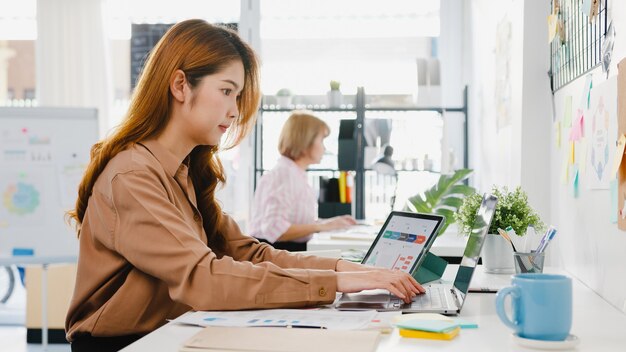 Junge Geschäftsfrau aus Asien, die soziale Distanzierung in einer neuen normalen Situation zur Virenprävention hält, während sie Laptop und Tablet wieder bei der Arbeit im Büro verwendet