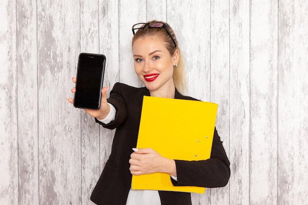 Junge Geschäftsfrau der Vorderansicht in der schwarzen Jacke der strengen Kleidung, die Dokumente und Telefon auf weißer Oberfläche hält