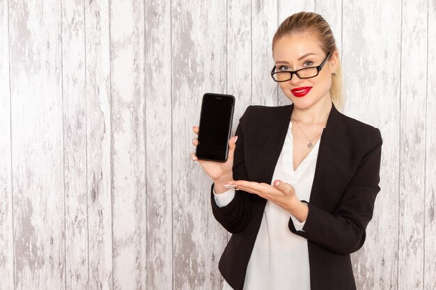 Junge Geschäftsfrau der Vorderansicht in der schwarzen Jacke der strengen Kleidung, die ihr Telefon auf weißem Schreibtisch hält